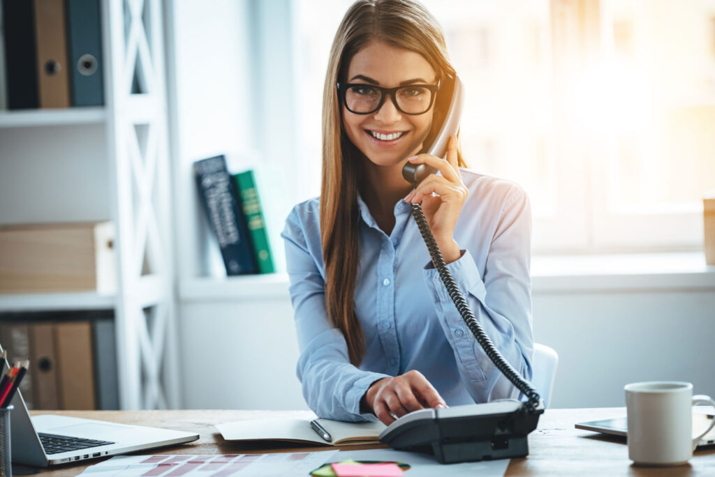 I will connect you in one second! Cheerful young beautiful woman in glasses talking on the phone and looking at camera with smile while sitting at her working place